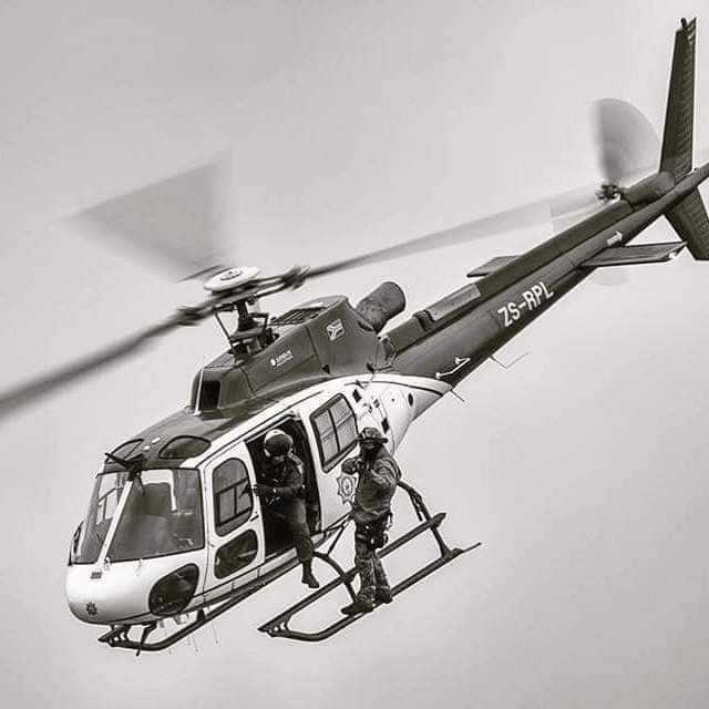 A helicopter in flight with two crew on the skids